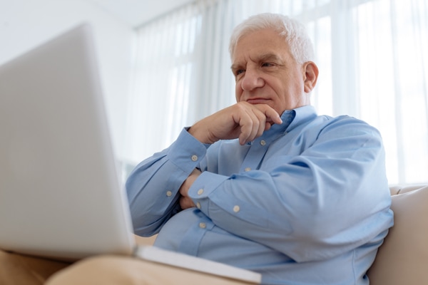 Elderly man sitting on comfy sofa and researching furnace sounds on laptop.