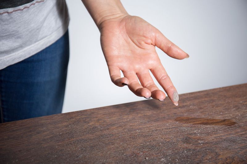 A hand with dust on the fingertips is above a table. How to Reduce Dust in Your Home.