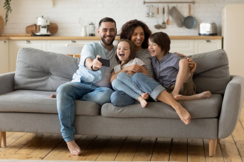 What Are the Signs that I Need a New Furnace? Photo of a family of four sitting on a couch together, laughing and posing for a photo on a phone.